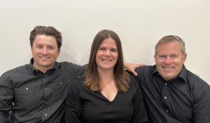 Portrait of a woman and two men in black professional attire, smiling in an office setting.
